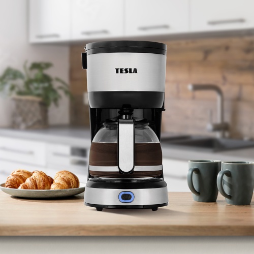 Predný pohľad na kávovar TESLA Coffeemaster ES200 s otvoreným vekom, vedľa pečivo a dva šálky na kuchynskej linke.