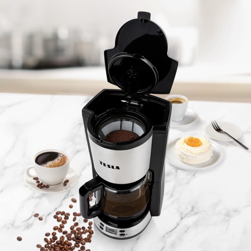 Otvorený kávovar TESLA Coffeemaster ES300 počas pridávania mletej kávy, s pripraveným dezertom a šálkou prekvapkávanej kávy na mramorovom pulte.